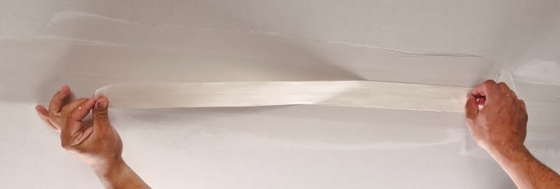Hände ziehen ein weißes Klebeband von einer Wand oder Decke ab.